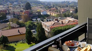 A balcony with a table set for breakfast, overlooking a town with buildings and mountains in the distance.