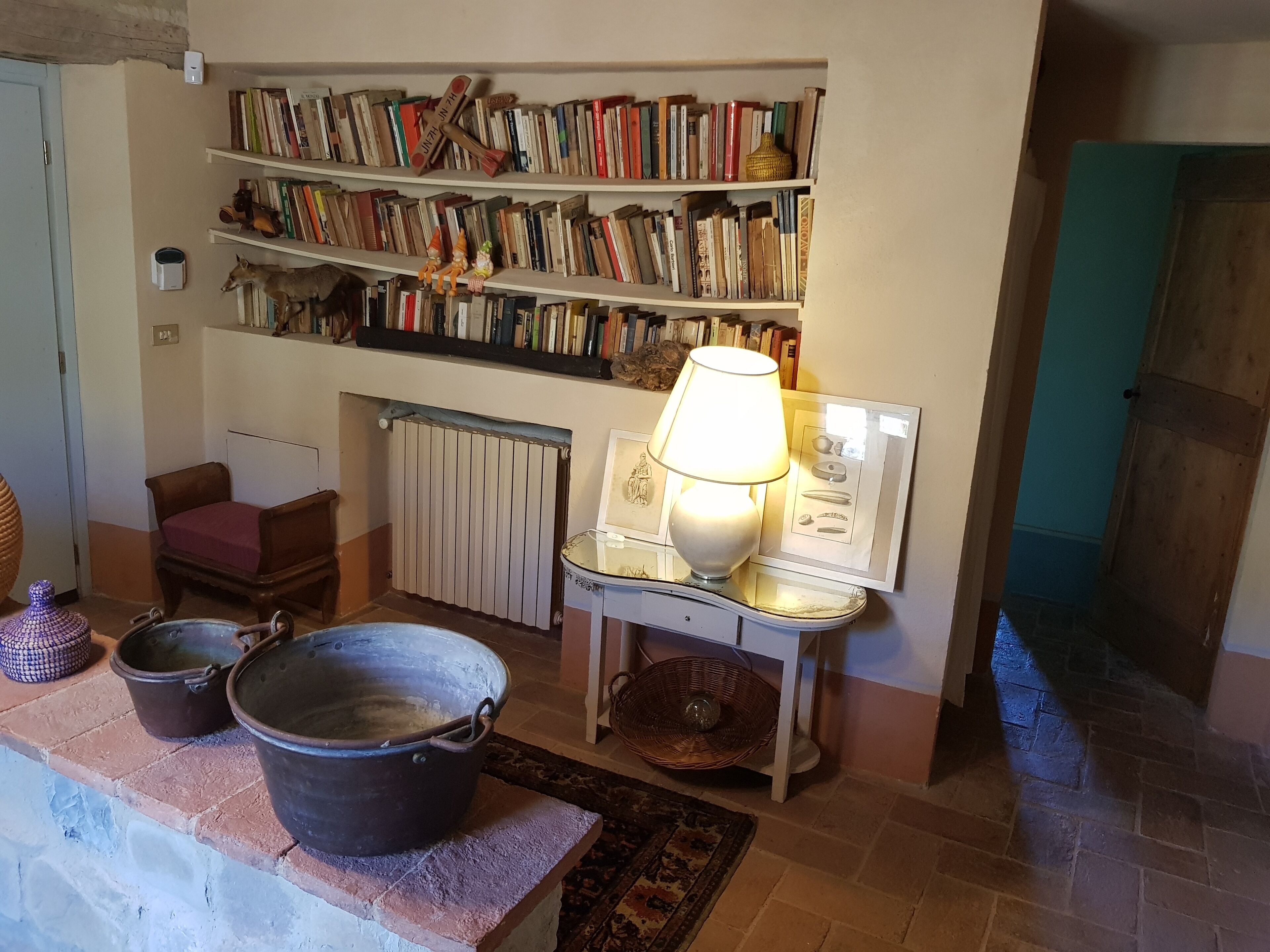 Een gezellige kamer met een open haard, een boekenplank vol boeken, een tafel met een lamp en twee grote potten op een plank.