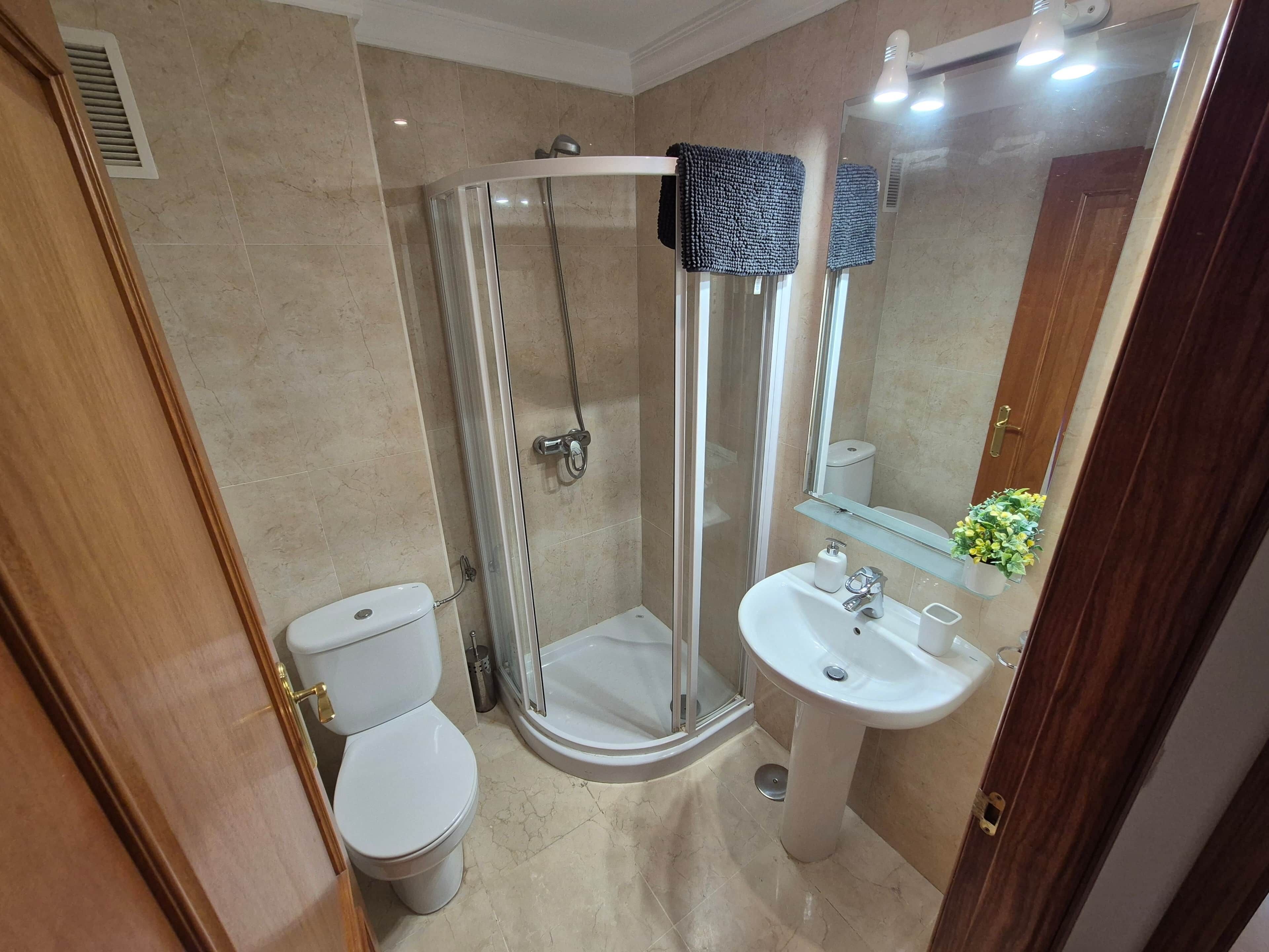 Un baño con inodoro, ducha con mampara de vidrio y lavamanos con espejo.