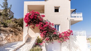 Um edifício de dois andares com varanda, escadaria de pedra e flores cor-de-rosa.