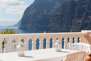 Ein weißer Tisch mit zwei weißen Stühlen und zwei weißen Laternen auf einem Balkon mit Blick auf das Meer und eine Klippe.