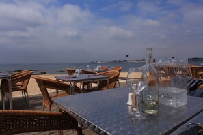 An outdoor dining area with wicker chairs and tables, overlooking a beach and the sea.