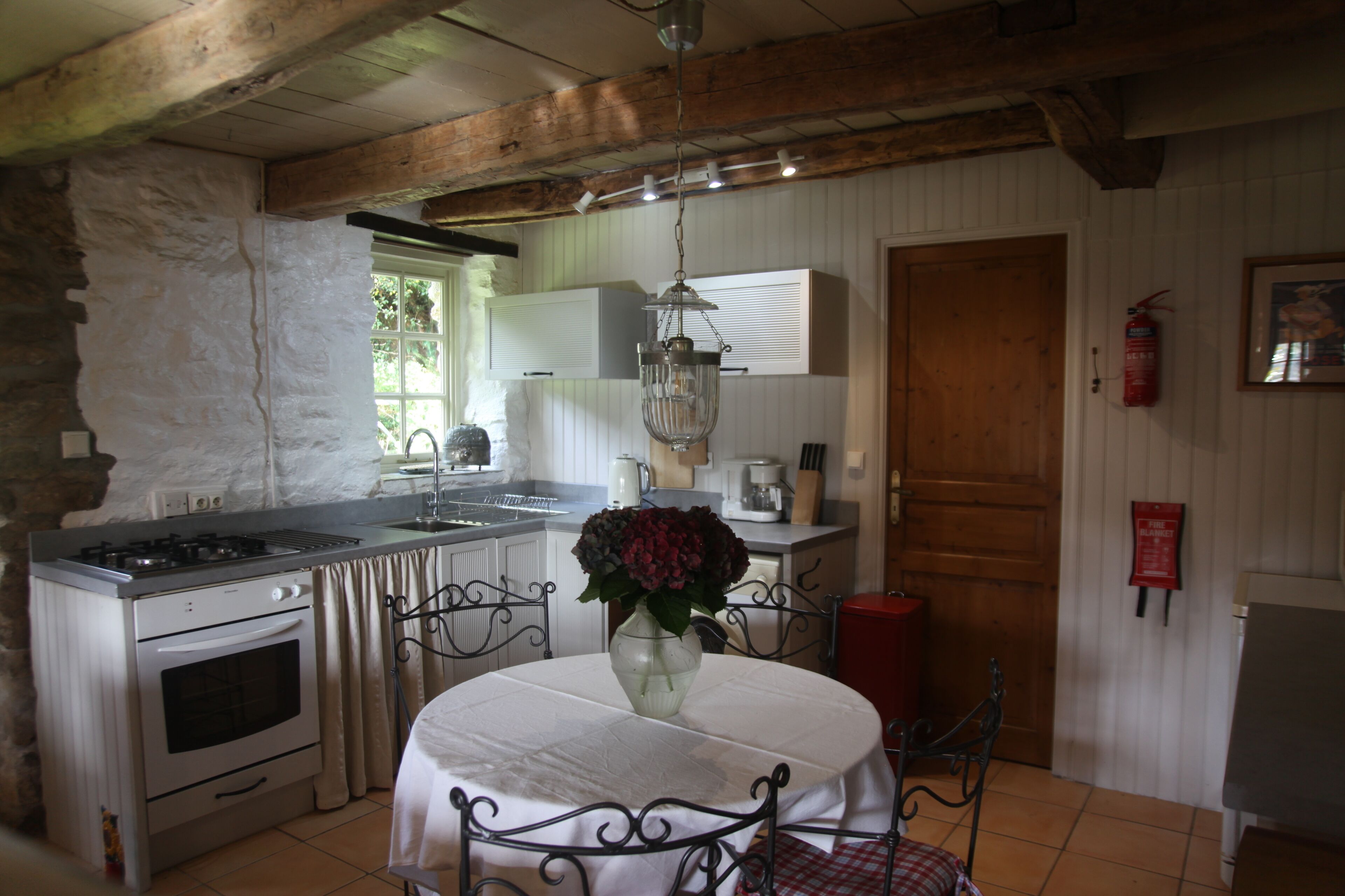 A rustic kitchen with a stone wall, wooden beams, a gas stove, an oven, a sink, and a dining table with chairs.