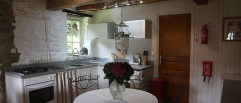 A rustic kitchen with a stone wall, wooden beams, a gas stove, an oven, a sink, and a dining table with chairs.
