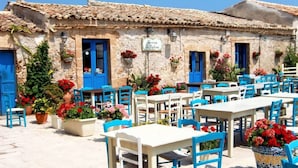 An outdoor café with white tables and blue chairs, surrounded by stone buildings with blue doors and flower pots. - House / Villa - Avola (Avola)