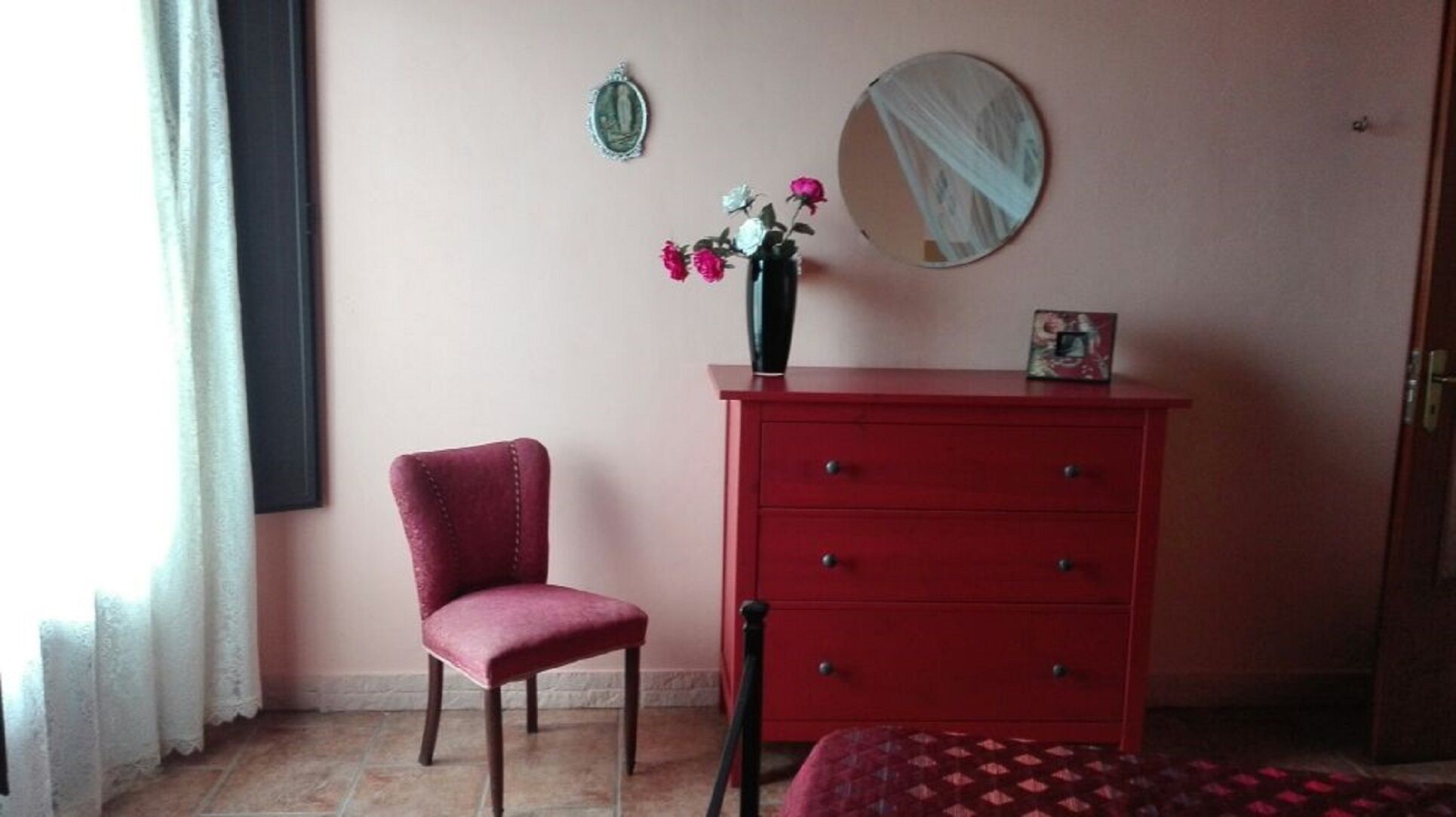 Ein Zimmer mit einem roten Schminktisch, einem Sessel, einem Spiegel und einer Vase mit Blumen.
