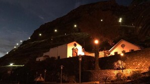 A building with a stone wall and a lit-up window, surrounded by a stone pathway and a palm tree. - Grannies House - Where Comfort and Relaxation Happens (Machico)
