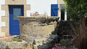Un bâtiment doté d’une façade en pierre, d’une porte bleue et d’une petite fenêtre. Un jardin avec différentes plantes et fleurs se trouve devant le bâtiment.