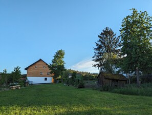Ein zweistöckiges Holzhaus mit geneigtem Dach, umgeben von einer Wiese und Bäumen.