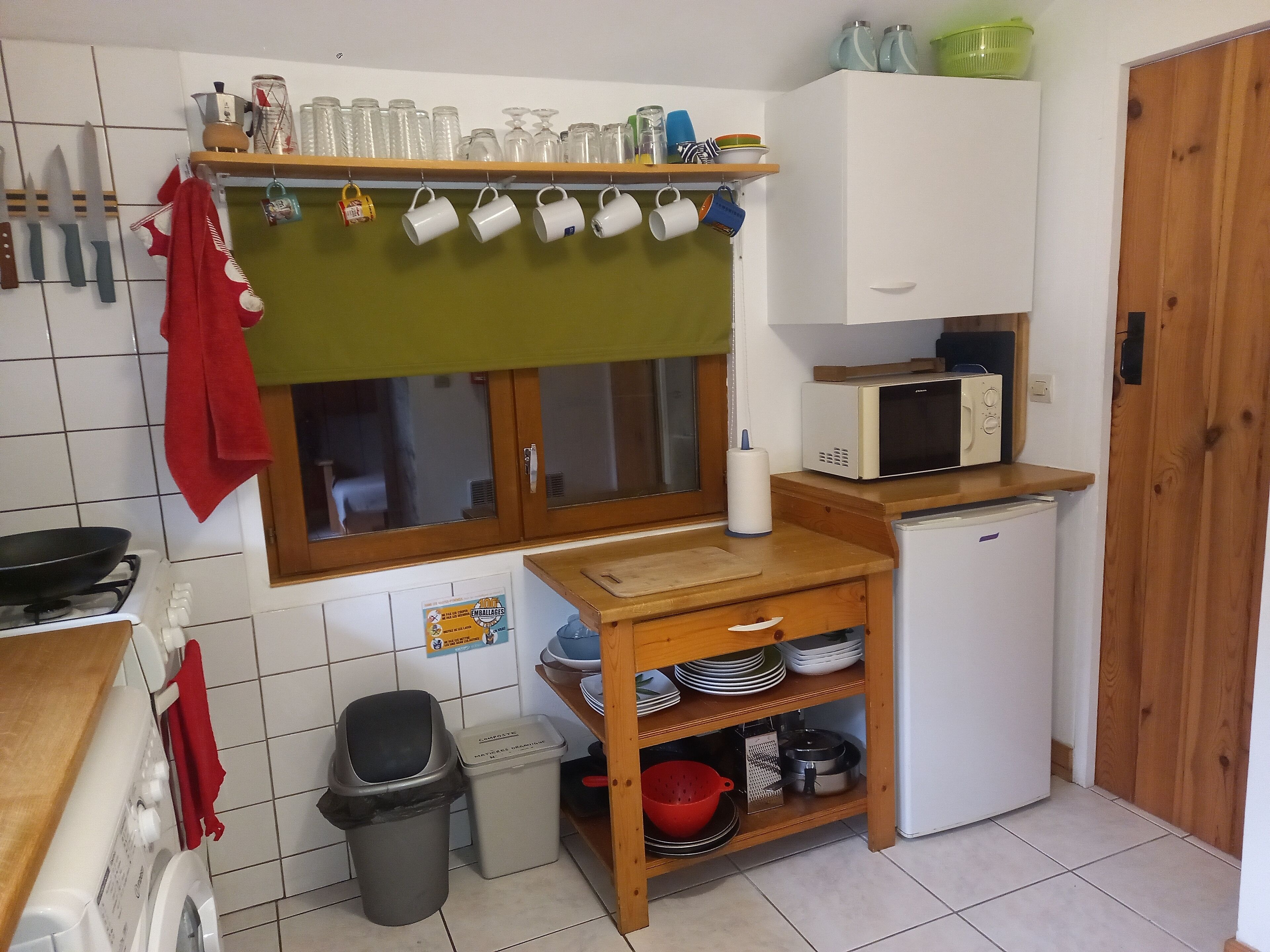 A compact kitchen with a microwave, a small refrigerator, and a wooden shelf with dishes.