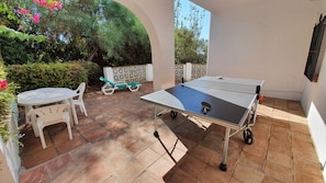 Mesa de ping pong con una raqueta, una silla, y una pequeña mesa con sillas en un espacio exterior cubierto con piso de baldosas.