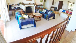 Una sala de estar con chimenea, sofá y mesa de centro. Se ve una estantería y un comedor al fondo.
