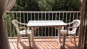 Een balkon met een witte tafel en twee stoelen, een witte reling, en uitzicht op bomen en huizen.