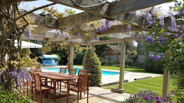 A wooden pergola with a pool table underneath, surrounded by lush greenery and purple flowers.