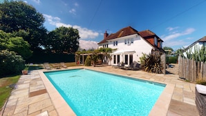 A rectangular swimming pool with a stone deck, surrounded by a white house with a red roof, a covered patio area, and a wooden fence.