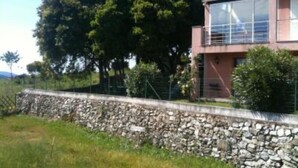 Uma casa moderna de dois andares com uma parede de pedra à frente, rodeada por vegetação e árvores.