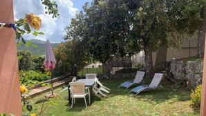 Um pátio externo com mesa e cadeiras, área coberta e vista para um muro de pedra e vegetação.