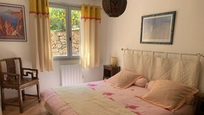 Quarto com cama, mesa de cabeceira, cadeira, janela com cortinas e um quadro na parede.