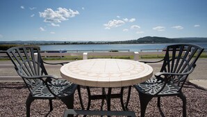 Uma mesa redonda com tampo de mosaico, rodeada por quatro cadeiras de metal preto, sobre um piso de cascalho com vista para um lago e colinas ao longe.