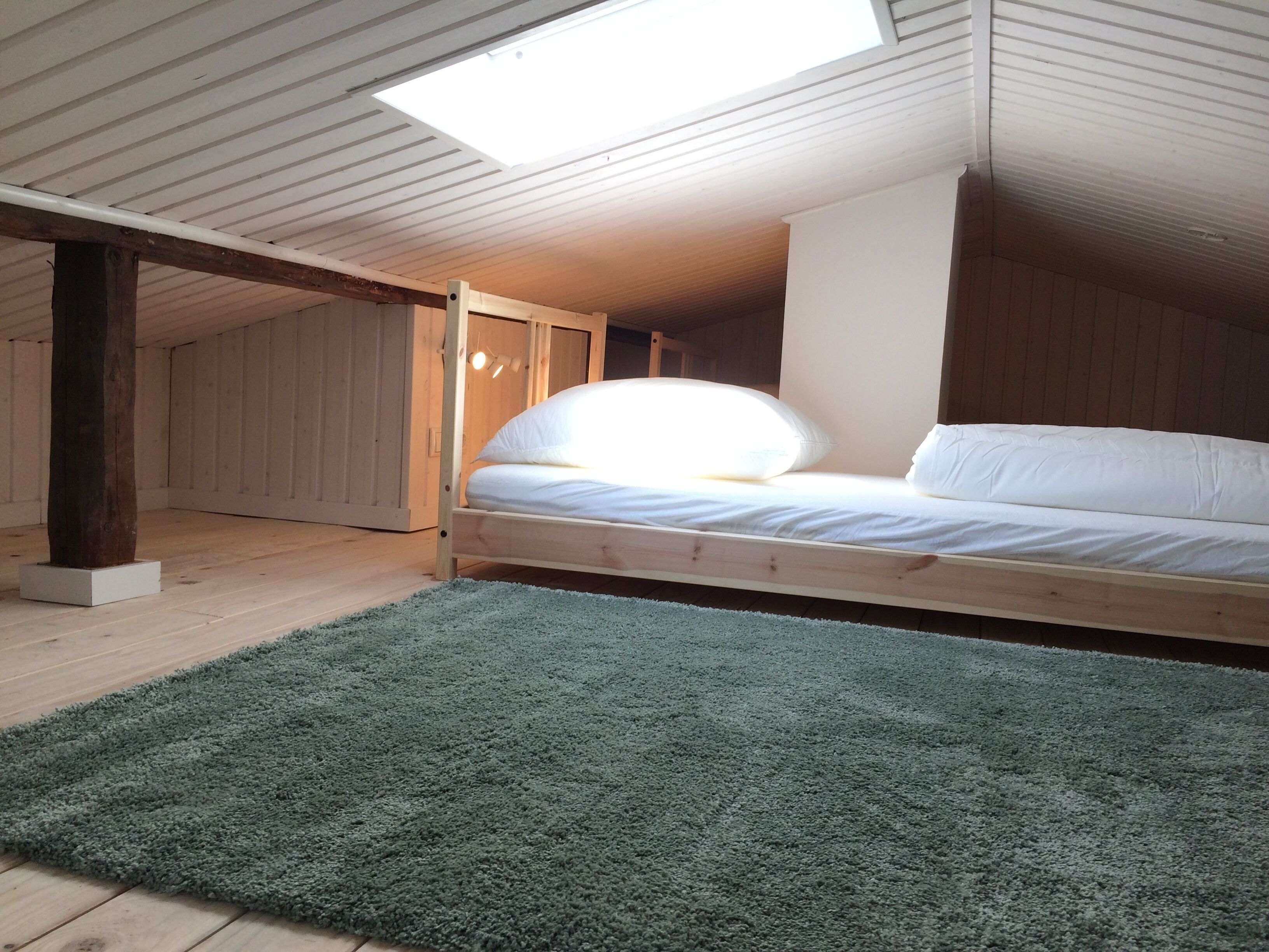 Ein Schlafzimmer im Loftsstil mit schrägem Dach, Holzbalken, einem Bett mit weißer Bettwäsche und einem Oberlicht.