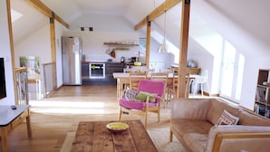 Ein geräumiger Wohnbereich mit einem Holztisch, einer Couch und einem Esstisch mit Stühlen. Im Hintergrund ist eine Küche zu sehen.