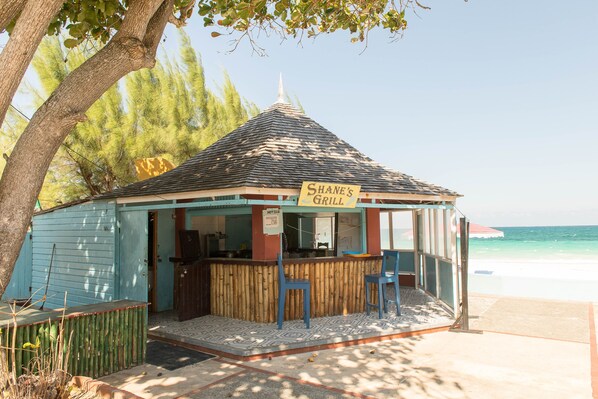 Una pequeña parrilla frente a la playa, con franjas azules y marrones, un letrero que dice "Shane's Grill" y dos sillas azules en el frente.