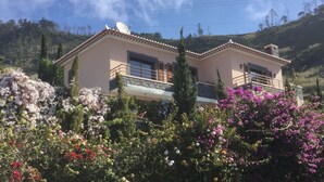 A two-story house with a balcony, surrounded by flowering plants and trees. -  Spacious villa,  180* ocean view, large heated pool, 5 min from Calheta beach (Calheta)