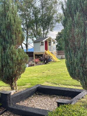 A small playhouse with a yellow slide, surrounded by a grassy yard and trees.