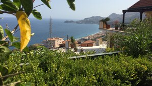 A coastal view with a building overlooking the sea, a wind turbine, and lush greenery.