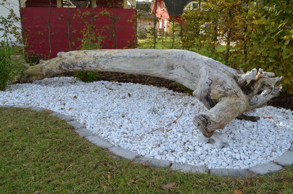 Une grande pièce de bois flotté, de forme courbée, placée dans un jardin, entourée de petits galets blancs et d’herbe.