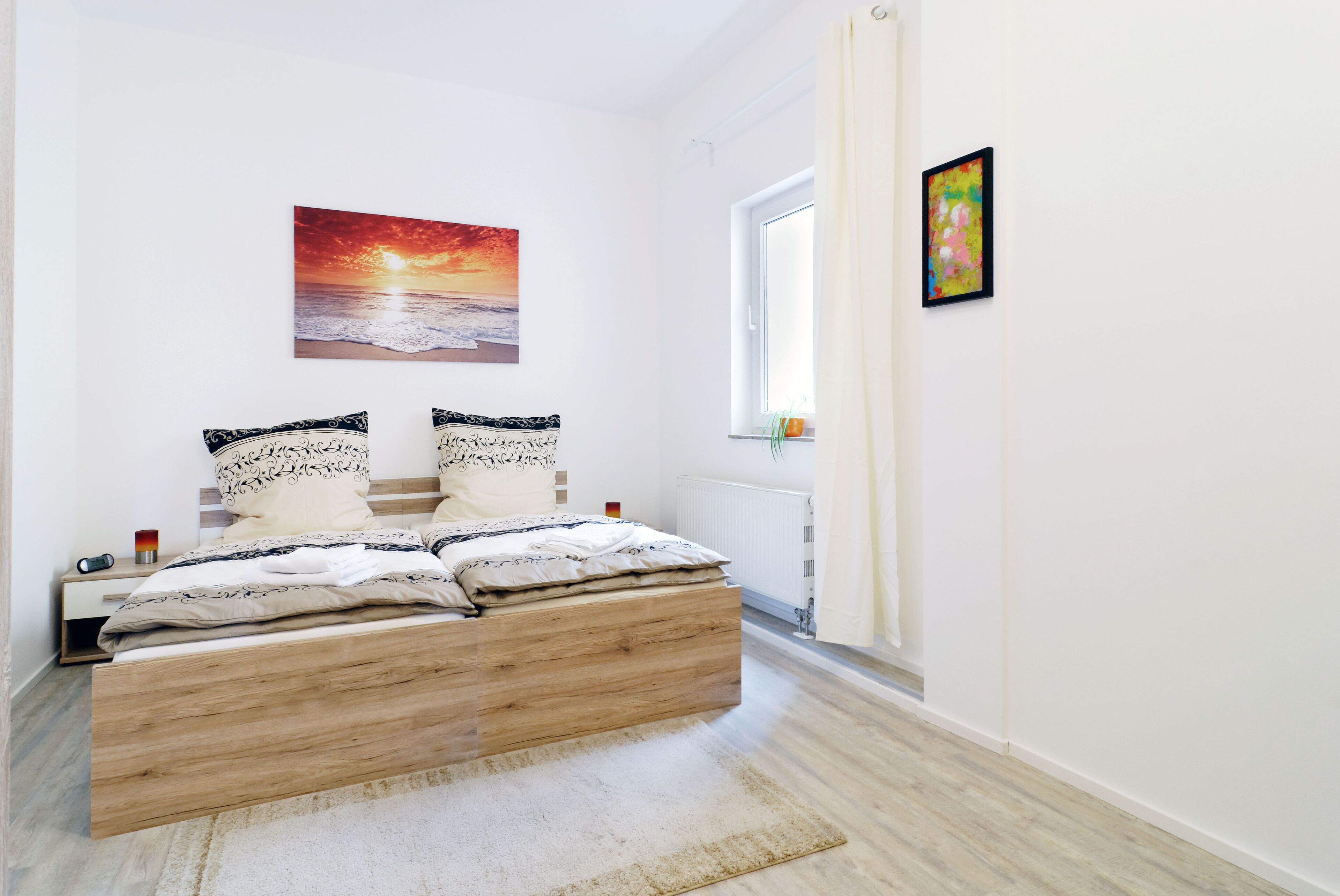 A bedroom with a wooden bed, a sunset painting, and a window with curtains.