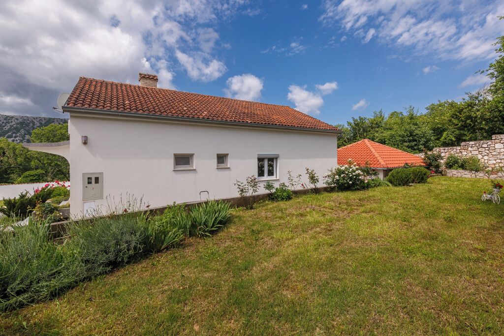 Une maison blanche à deux étages, avec un toit recouvert de tuiles rouges, entourée d’une pelouse verdoyante et d’un mur de pierres.