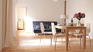 Un salon moderne avec un plancher en bois, un canapé noir, des chaises blanches, une table à manger en bois et un vase rempli de fleurs.