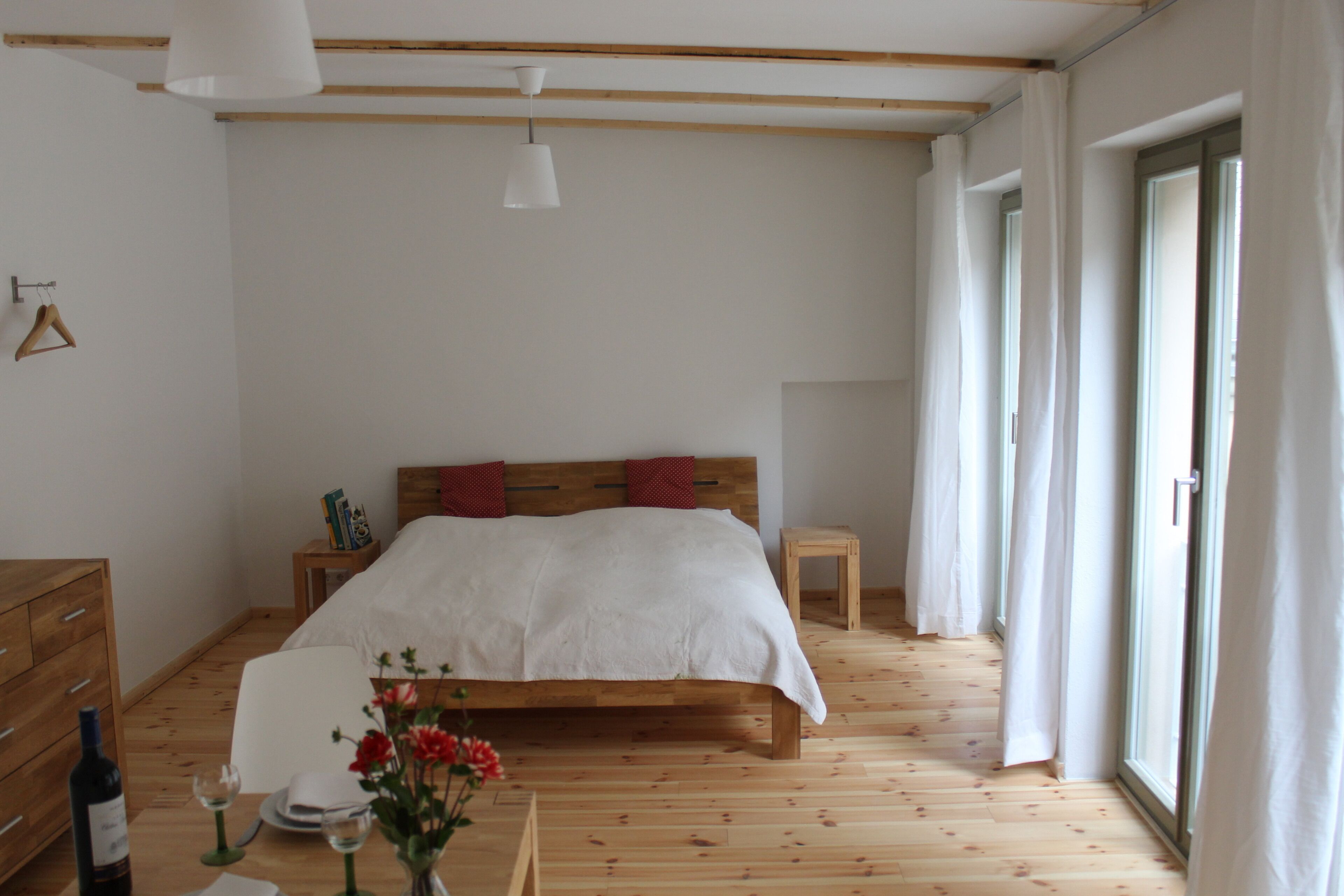 Un dormitorio con una cama de madera, una mesita de noche, una pequeña mesa con una botella de vino y copas, y una ventana con cortinas.