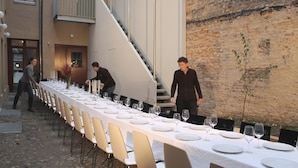 Une longue table à manger, recouverte de nappes blanches, entourée de chaises, avec un mur en briques et un escalier.