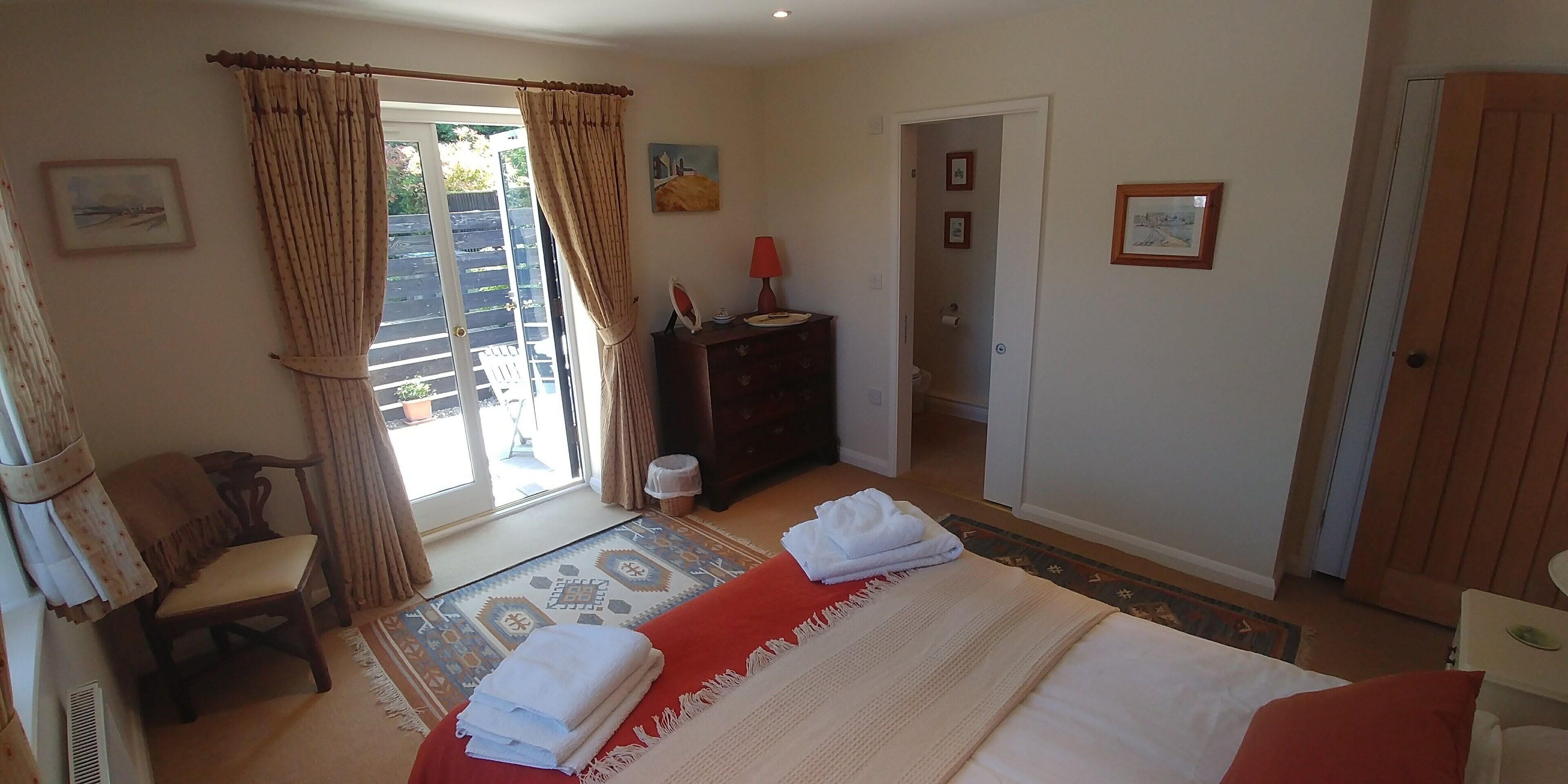 A hotel room with a bed, a chair, a small table with a lamp, a wardrobe, and a door leading to a balcony.