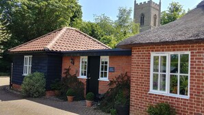 A traditional brick building with a tiled roof, a sign reading "FOTTS COTTAGE," and a church tower in the background. - Country Cottage Set In pretty Suffolk village with 2 local pubs, dog friendly (Lower Ufford)