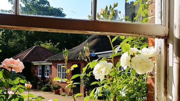 A view through a window of a brick house with a thatched roof, surrounded by lush greenery and blooming flowers. - Country Cottage Set In pretty Suffolk village with 2 local pubs, dog friendly (Lower Ufford)