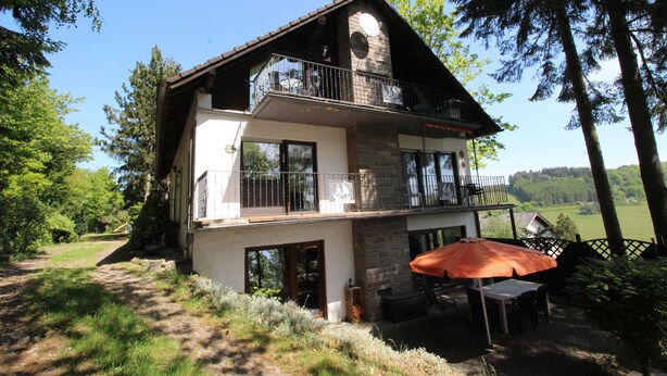 A two-story house with a balcony, a patio with an umbrella, and a view of the surrounding landscape. - Cozy, comfortably furnished apartment for up to 7 people with balcony (Immerath)