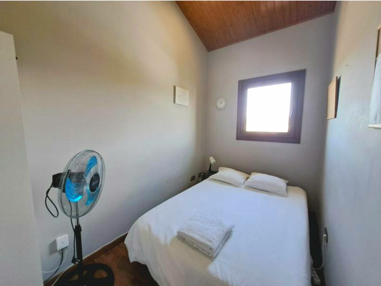 A bedroom with a bed, a standing fan, a window, and a clock on the wall.