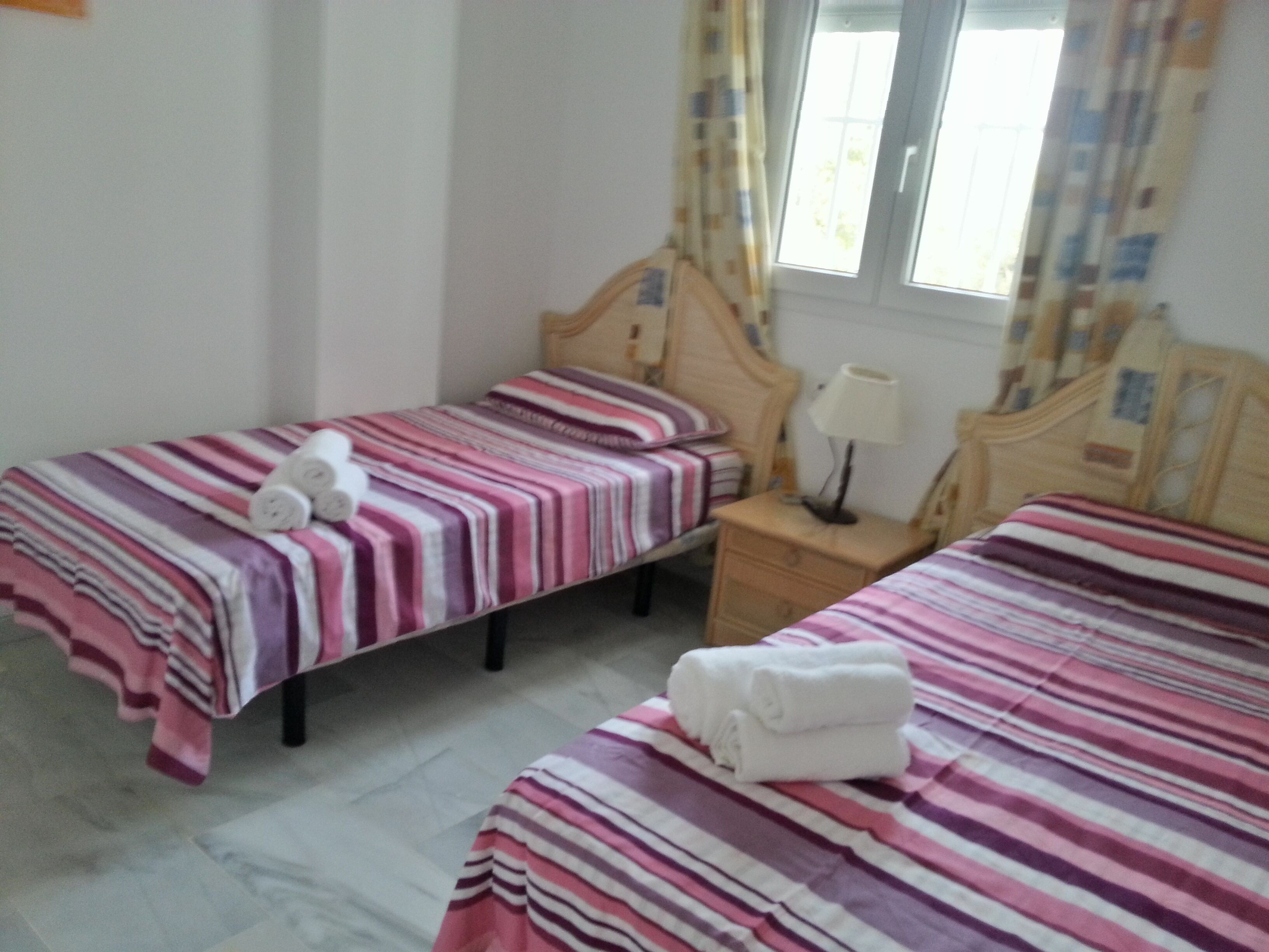 A hotel room with two single beds, each with a striped bedspread and a rolled towel. There is a wooden nightstand with a lamp between the beds.