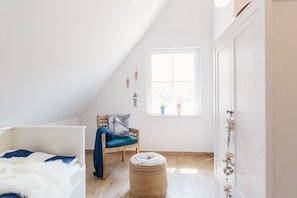 Quarto com teto inclinado, cama com cabeceira branca, cadeira azul com almofada, um pequeno banquinho e janela com vista para o exterior.