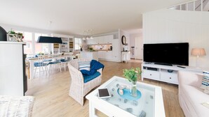 A modern living room with a glass coffee table, a television, and a dining area with white chairs and a blue cushion.