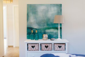 Um console branco com um quadro abstrato azul-esverdeado, duas garrafas verdes, uma tigela decorativa, um abajur e três cestos de vime com recortes em formato de coração.