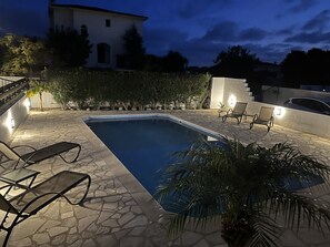A swimming pool with lounge chairs and a stone pathway, surrounded by a hedge and a white building in the background.