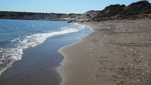 Una platja amb cel blau, mar tranquil·la i penya-segats rocosos al fons.