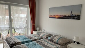 A bedroom with a bed, a nightstand, a lamp, a painting of a lighthouse, and a view of the outdoors through a window with curtains. - Great apartment with good facilities in a prime location in Schillig (Wangerland)