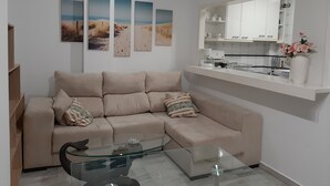 A living room with a beige sofa, a glass coffee table, and a kitchen area with a white countertop and cabinets. - SPECIAL FAMILIES, WI-FI, BEACH, POOL, SPORTS TRACKS (Málaga, Benalmádena)