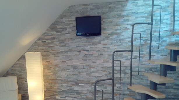 A modern interior with a stone-patterned wall, a flat-screen TV mounted above a staircase, and a white chair. - in a good location to Dusseldorf and Cologne and the JEVER Skihalle Neuss (Grevenbroich)