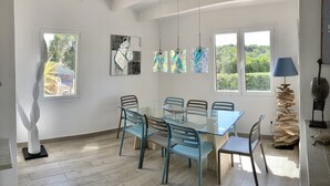 A dining area with a glass table and blue chairs, surrounded by windows with a view of greenery. - Stylish, spacious and airy, in a quiet area  but close to beach, restos & shops (Sant Lluís)